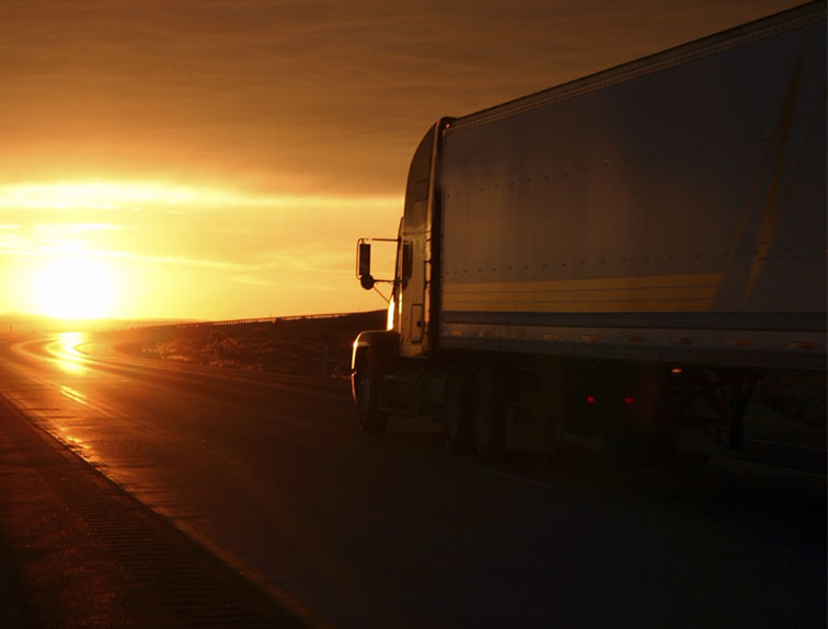 Image of a semi truck driving heading toward a sunset.