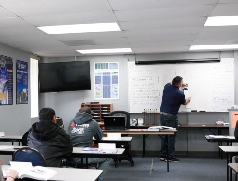 A CDL instructor writes a document checklist on a whiteboard while students take notes in a classroom