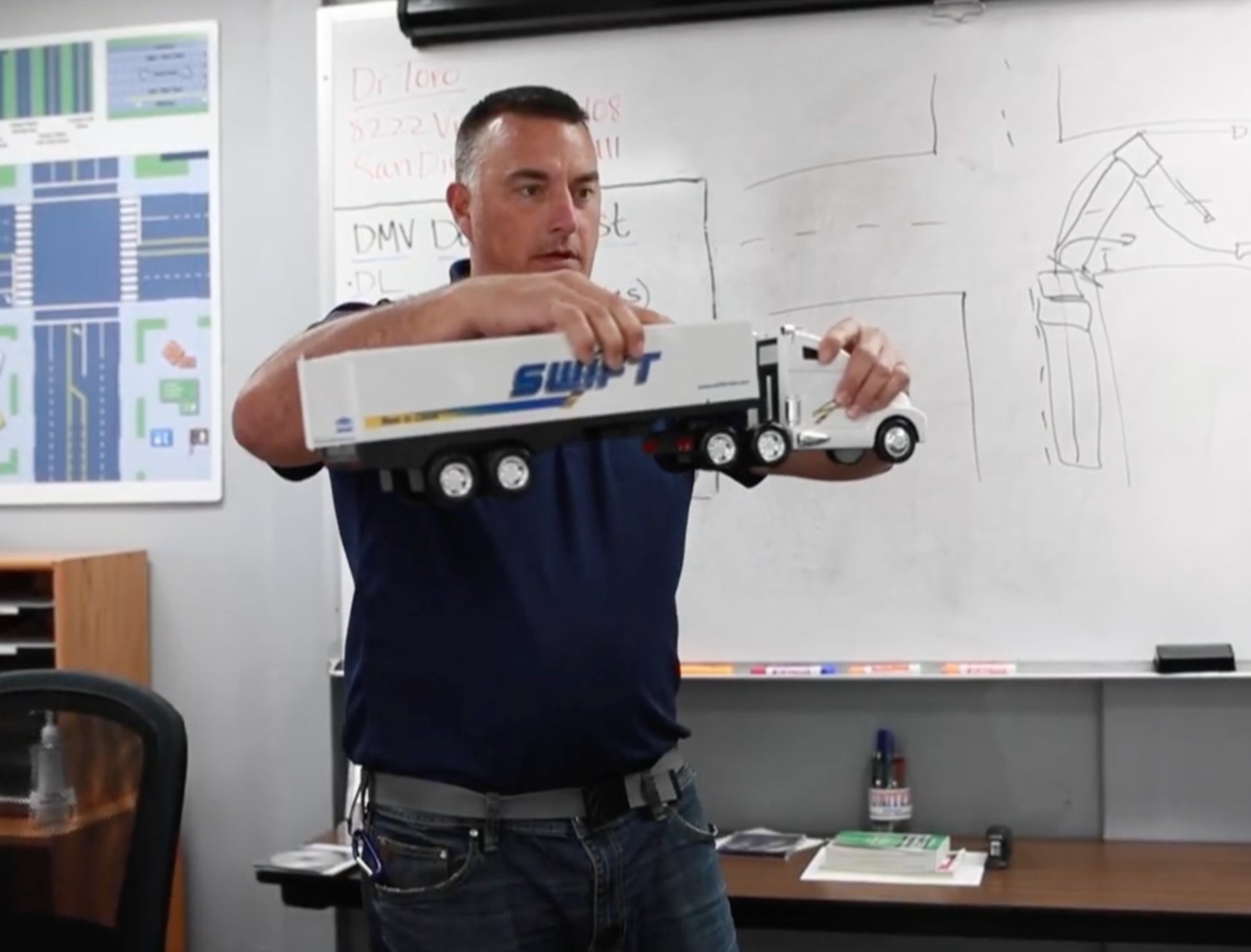 A CDL instructor in a classroom holding a model Swift semi-truck to demonstrate turning maneuvers, with a whiteboard and road diagrams behind him.