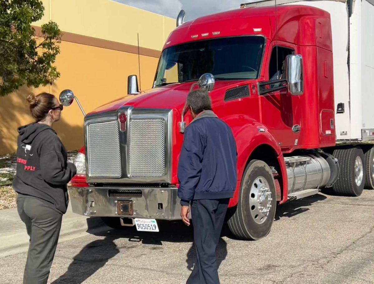A CDL instructor and student standing beside a red semi-truck during a pre-trip inspection in an outdoor lot.